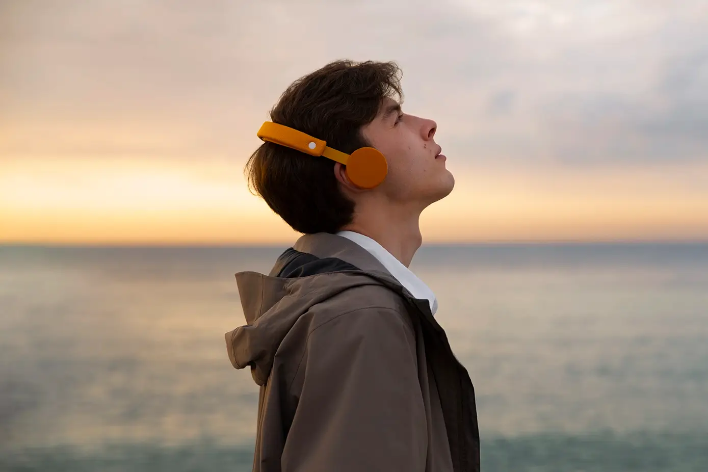Jovem de perfil usando fones de ouvido laranja e olhando para o céu ao entardecer à beira-mar, representando a introspecção necessária para que a IA não enferruje o cérebro.