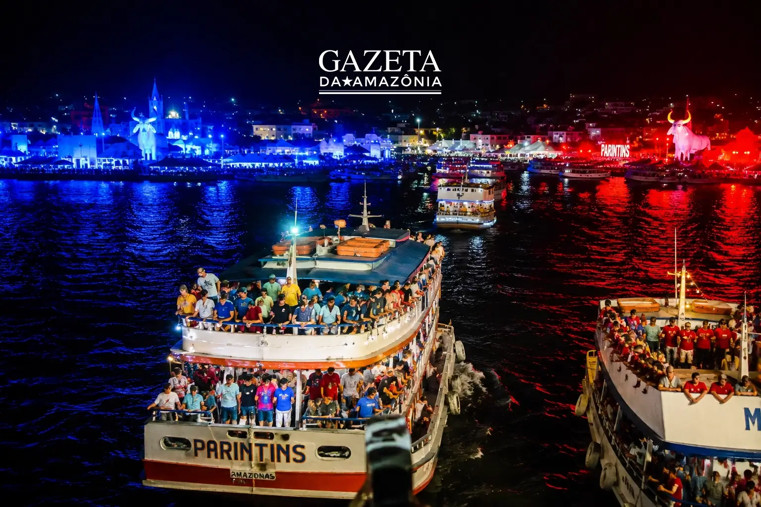 Vista aérea noturna de embarcações repletas de passageiros navegando em direção à ilha durante o Festival de Parintins 2026, mostrando a orla iluminada com as cores azul do Caprichoso e vermelha do Garantido.